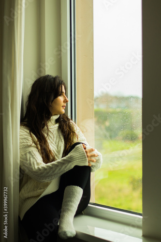 Mujer sentada en la ventana con lluvia mirando mirando hacia el exterior pensativa con paisaje verde