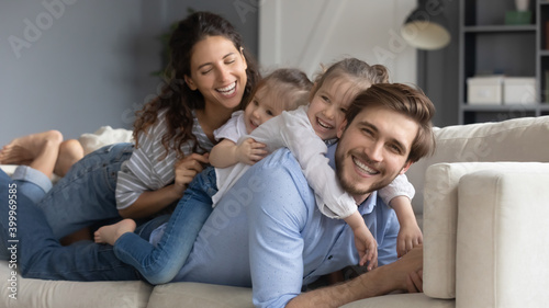 Wide banner panoramic portrait of happy young Caucasian family with two small daughters relax on couch at home. Smiling parents rest on sofa in living room together with little girls children.