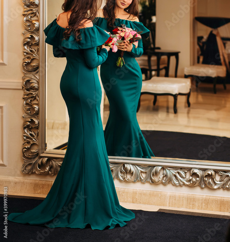 Mysterious Young beautiful stylish woman in green emerald evening dress, silhouette of a mermaid stands looking at herself in mirror, holding a bouquet of flowers in her hands. classic luxury interior