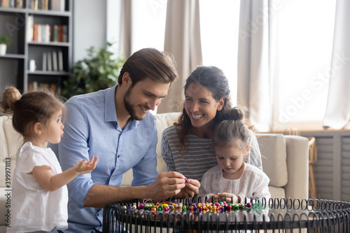 Overjoyed young Caucasian family with two small daughters feel playful making bracelets together at home. Happy loving parents have fun play string colorful beads on thread with small girl children.