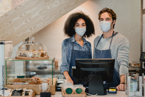 Two cafeteria staff standing at cafe counter in medical masks against Covid 19 in small coffee shop