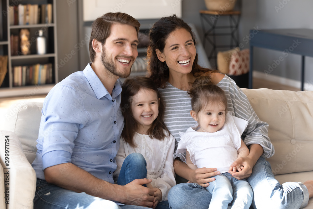 Overjoyed young Caucasian mom and dad sit rest on sofa with two little daughters have fun on weekend at home together. Happy family with small girls children relax on couch playing laughing.
