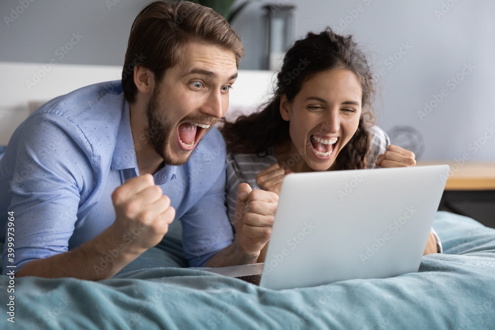 Overjoyed millennial man and woman lying in bed scream celebrate online victory or win on laptop. Excited young Caucasian couple feel euphoric triumph with good news on computer. Success concept. Overjoyed millennial man and woman lying in bed scream celebrate online victory or win on laptop. Excited young Caucasian couple feel euphoric triumph with good news on computer. Success concept.