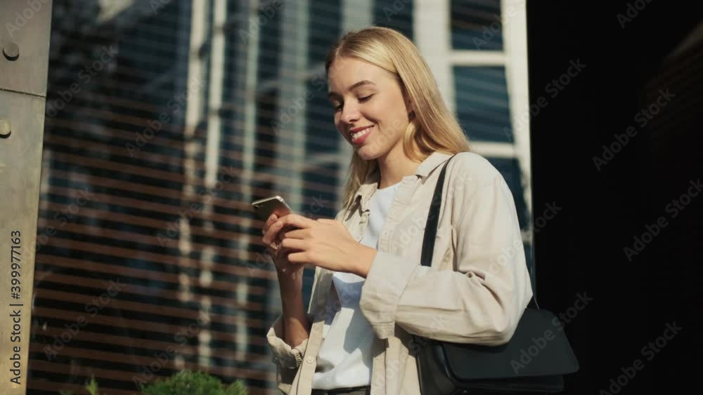 A focused young blonde woman is using her smartphone standing outside in the street near the building