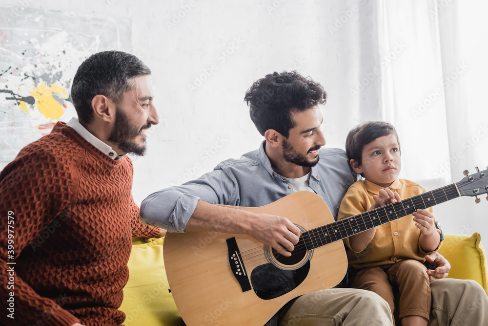 Fototapeta premium Smiling hispanic father playing acoustic guitar near son and grandfather at home, three generations of men