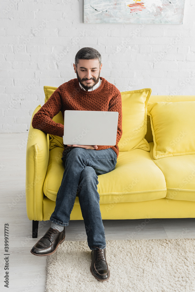Mature hispanic man using laptop while sitting with crossed legs on couch in living room