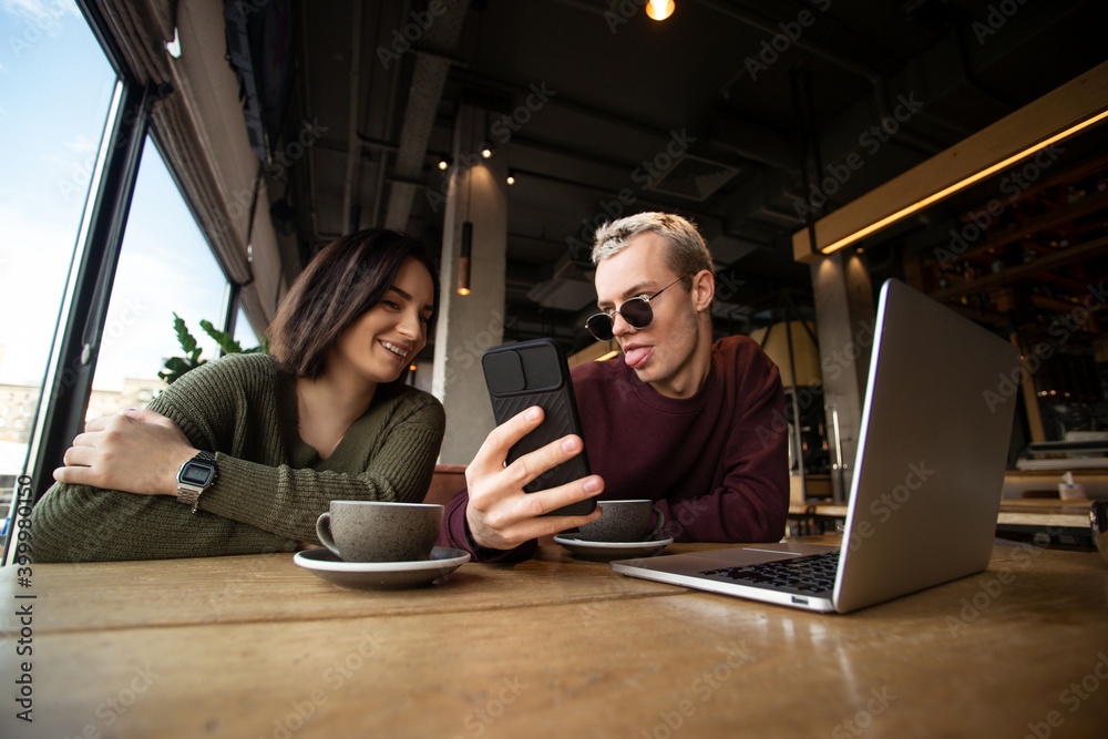 Man showing memes to woman. Lunch break concept. Young attractive man showing tongue and funny message on his smartphone to beautiful brunette woman. Working in coffee shop concept. Man showing memes to woman. Lunch break concept. Young attractive man showing tongue and funny message on his smartphone to beautiful brunette woman. Working in coffee shop concept.