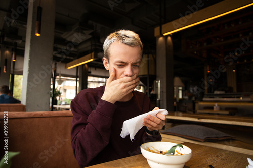 Spoiled food in the restaurant. Dissatisfied young man in a cafe covers his mouth with a hand disgusted with a taste of food. A meal on the table in front of him. Bad customer service concept.
