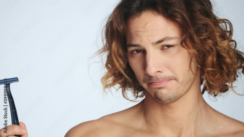 A confused man is holding a razor standing isolated over white wall in studio A confused man is holding a razor standing isolated over white wall in studio