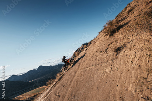 Extreme Rider climbing sand mountain top on off-road cross enduro motorcycle. Beautiful mountains landscape down on background, colourful autumn forest and river in sunshine