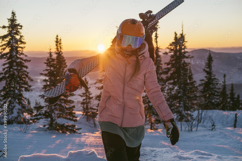 Skier female holding ski standing on mountain top during sunset, beautiful winter mountains landscape