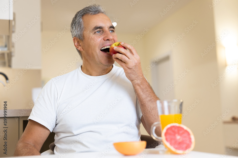Happy adult man eating fruits and healthy food on breakfast. Morning routine concept. Cut halves of grapefruit and glass of orange juice on table. Cozy modern kitchen on background. Happy adult man eating fruits and healthy food on breakfast. Morning routine concept. Cut halves of grapefruit and glass of orange juice on table. Cozy modern kitchen on background.