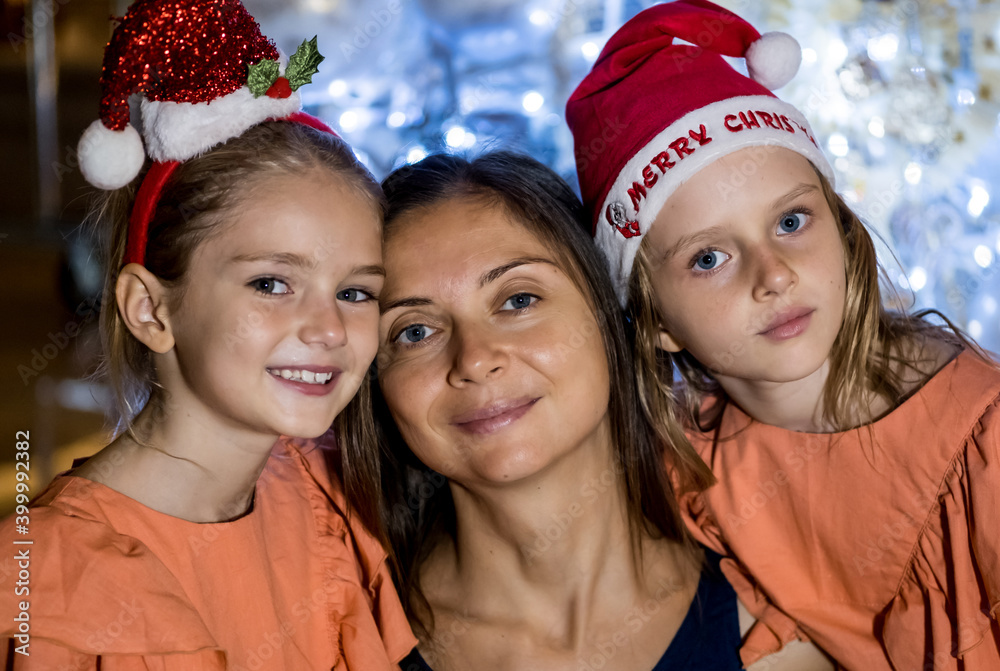 Single mother and two daughters by the Christmas tree wearing festive hats. Merry Christmas and happy holidays Single mother and two daughters by the Christmas tree wearing festive hats. Merry Christmas and happy holidays