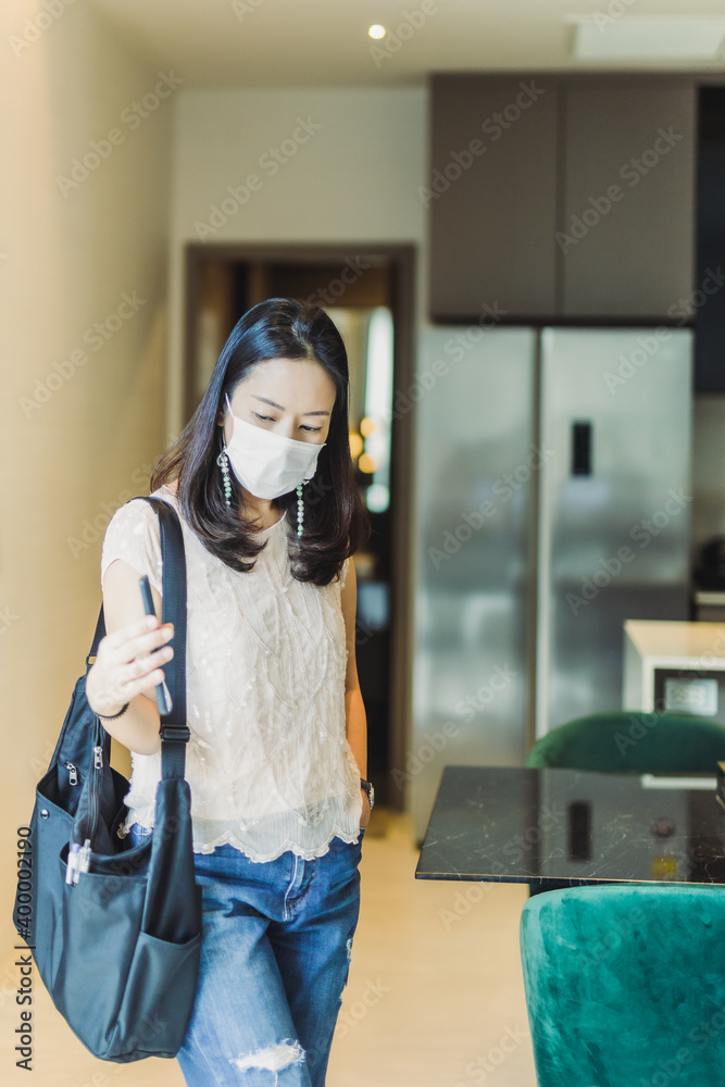 Portrait woman wearing protective mask holding mobile phone while standing in kitchen at home.