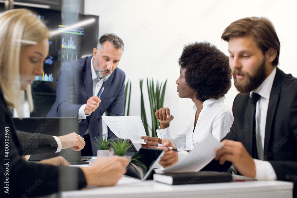 Confident middle aged male boss talking and cooperating with diverse focused business team at office briefing, discussing business ideas and showing smth on paper to african woman Confident middle aged male boss talking and cooperating with diverse focused business team at office briefing, discussing business ideas and showing smth on paper to african woman