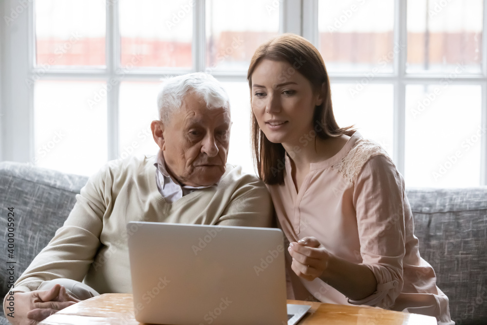 Young female social worker volunteer help assist retired elderly man in shopping online using home computer. Focused old father look at laptop screen watch grown daughter teaching him pay bills on pc Young female social worker volunteer help assist retired elderly man in shopping online using home computer. Focused old father look at laptop screen watch grown daughter teaching him pay bills on pc