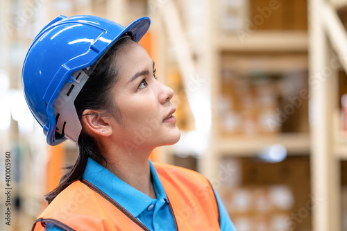 Close up woman worker standing in the automotive parts warehouse store and look on the shelf. Female engineers wear a safety helmet, vest standing in the factory. In background shelves with goods