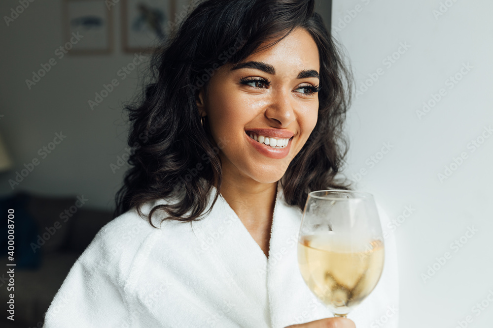 Beautiful cheerful woman with bocal of wine standing in living room. Young female relaxing at home alone. Beautiful cheerful woman with bocal of wine standing in living room. Young female relaxing at home alone.