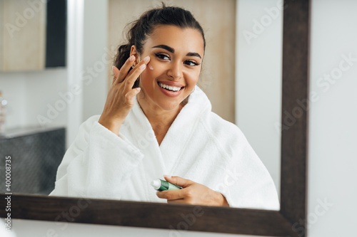 Middle east woman applying cream in bathroom mirror. Young smiling female putting moisturizer on her face.