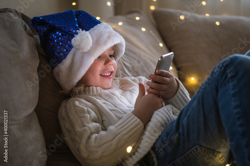 Girl in blue Santa hat using mobile phone for video call friends and grandparents.