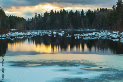 Frosty winter lake. Nothern snowy landscape. Stones and ice. Sunset and reflections on water surface. Panoramic view
