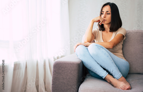 Tired and sad young woman is sitting and thinking on the couch at home and looking on the window