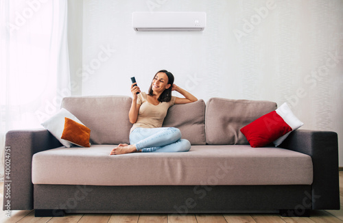 Beautiful modern brunette woman is using the air conditioner while sitting on the couch and resting in the hot day photo