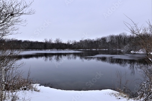 Winter landscape, lake among forest trees, trees are reflected in the water. There is snow on the ground, but the water in the lake is not frozen yet