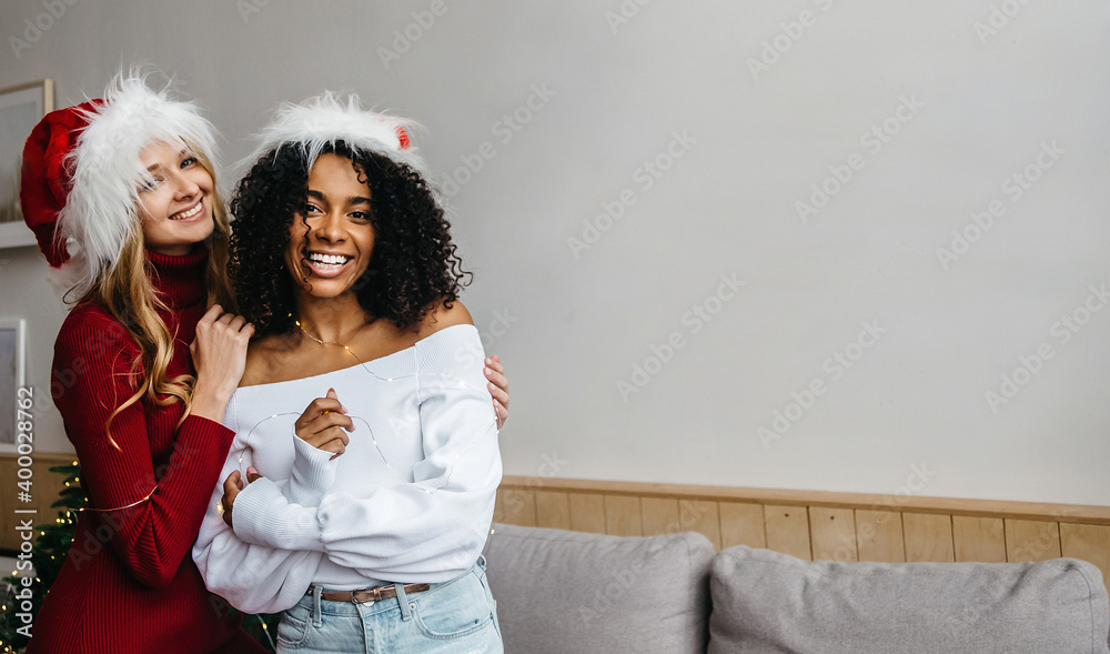 Fototapeta premium Christmas unites racial diversity. African and caucasian young women with Santa hat at home hugs and looking at camera. Black lives matter and tolerance concept