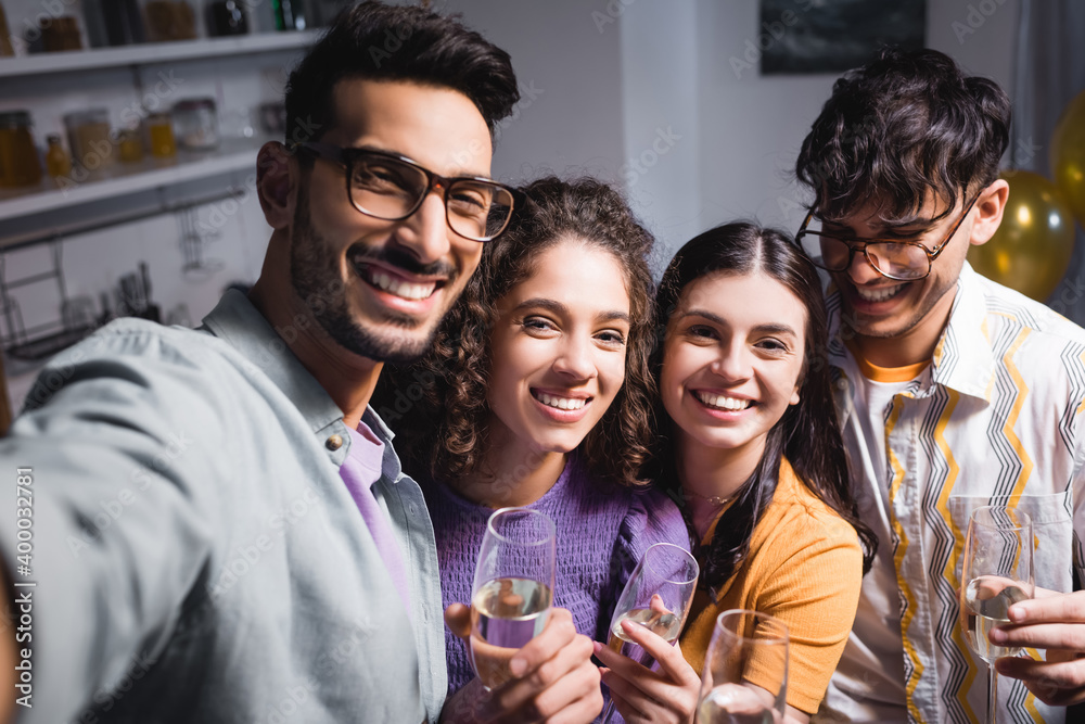 Obraz premium happy, young hispanic friends smiling at camera while holding champagne glasses