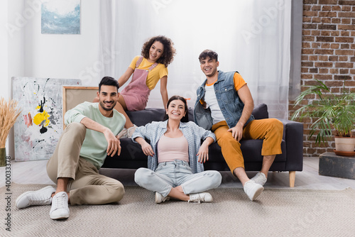 happy hispanic friends looking at camera while sitting on sofa and floor in living room