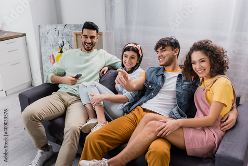 smiling man pointing with finger while watching tv with cheerful hispanic friends