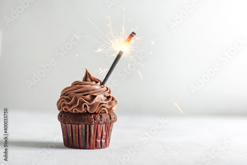 Cupcakes or muffins with chocolate cream and sparklers on light marble background. Holiday cake celebration, delicious dessert, close up. Copy space