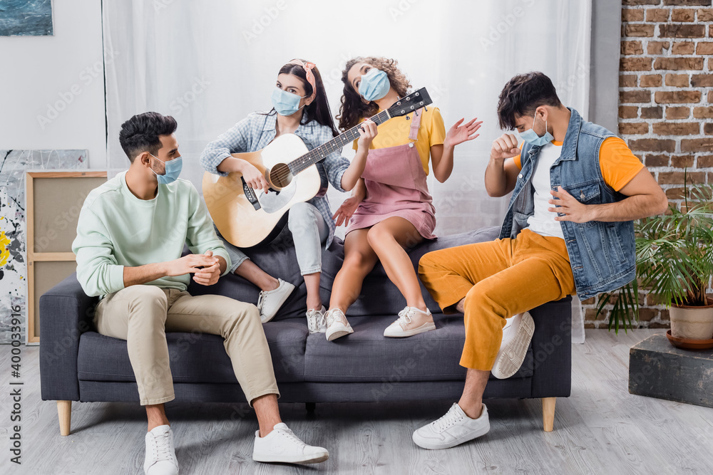 Hispanic friends in medical masks playing acoustic guitar on sofa in living room Hispanic friends in medical masks playing acoustic guitar on sofa in living room