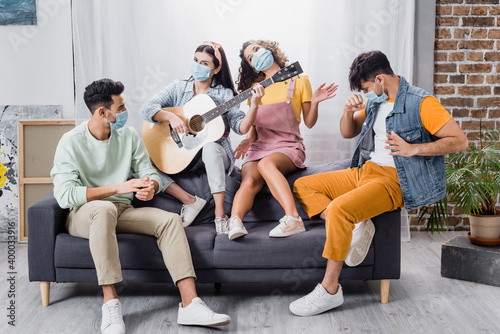 Hispanic friends in medical masks playing acoustic guitar on sofa in living room