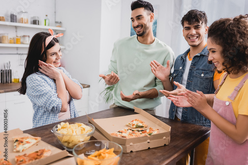 Smiling hispanic friends applauding near pizza and potato chips on blurred foreground in kitchen