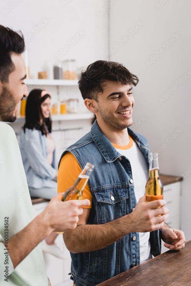 Smiling hispanic man holding bottle of beer near friend on blurred foreground in kitchen Smiling hispanic man holding bottle of beer near friend on blurred foreground in kitchen