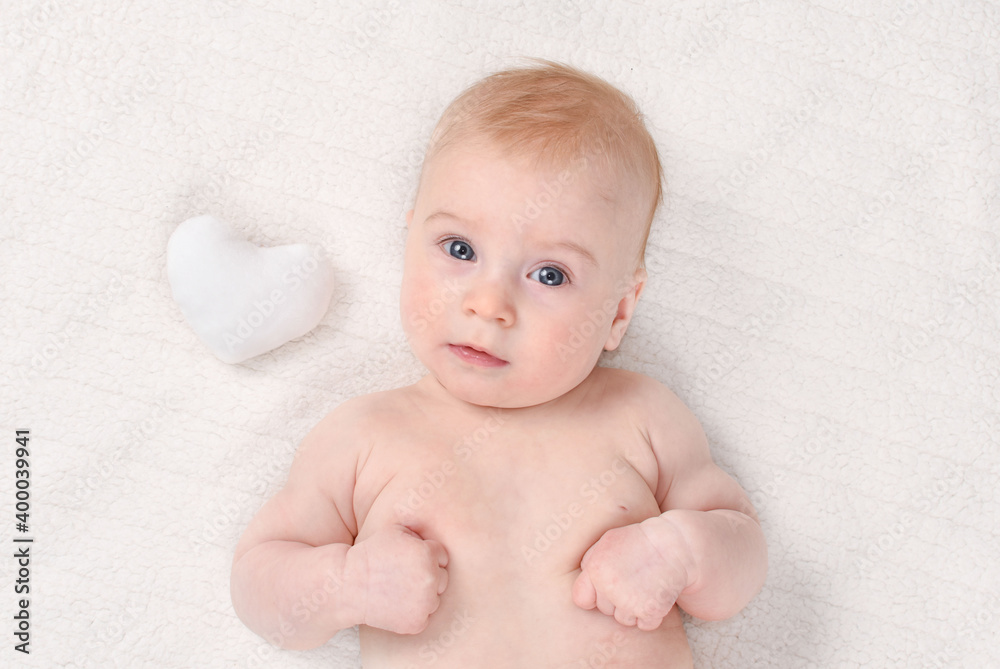 Cutest little 4- 5 months old caucasian baby girl with white soft heart in striped T-shirt on white plaid. Valentine day concept. Look at camera. Cutest little 4- 5 months old caucasian baby girl with white soft heart in striped T-shirt on white plaid. Valentine day concept. Look at camera.