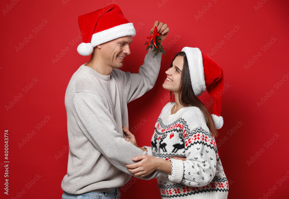 Obraz premium Happy couple in Santa hats standing under mistletoe bunch on red background