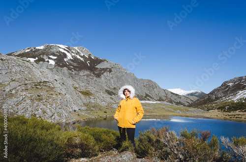 Mountain landscape with snowy mountains and lake. Woman in yellow jacket looking at camera.