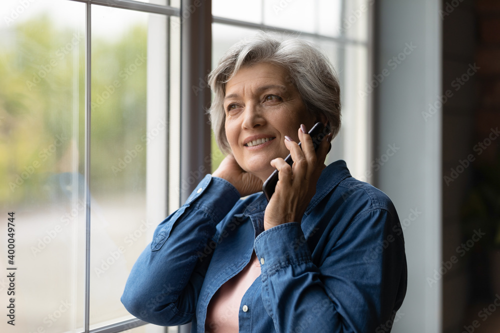 Happy middle aged 50s woman standing near window, enjoying pleasant mobile phone conversation with friends. Smiling dreamy older mature lady communicating distantly by smartphone call indoors. - obrazy, fototapety, plakaty 
