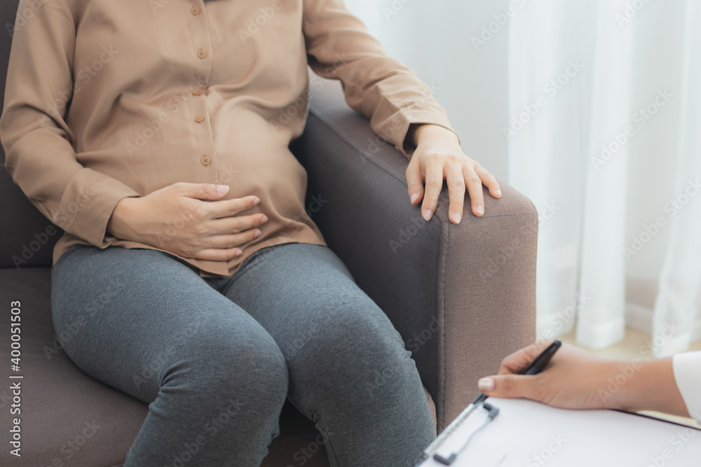 Young asian pregnant woman holding her belly while gynecologist notes the symptoms that the pregnancy is explaining about the unborn child.