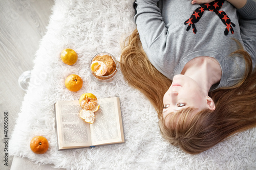 Young woman lying on bed with book and mandarins on white fur bedspread in Christmas sweater, festive atmosphere, Christmas atmosphere. Pleasure, rest, relaxation, dreams, make a wish. flat lay