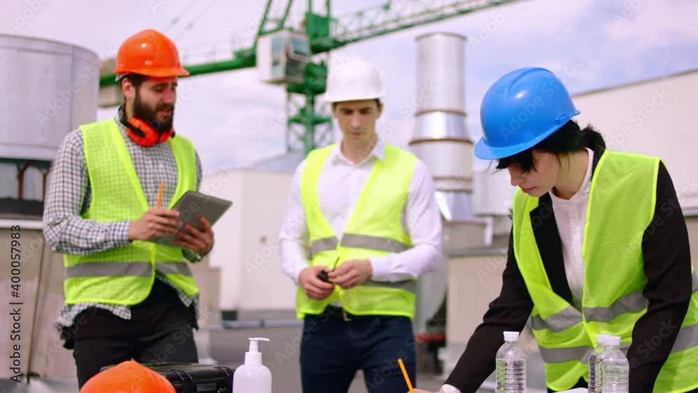 The construction worker one lady engineer and architect all together meeting on the roof top of construction site and analyzing the plan of construction they wearing safety helmets The construction worker one lady engineer and architect all together meeting on the roof top of construction site and analyzing the plan of construction they wearing safety helmets