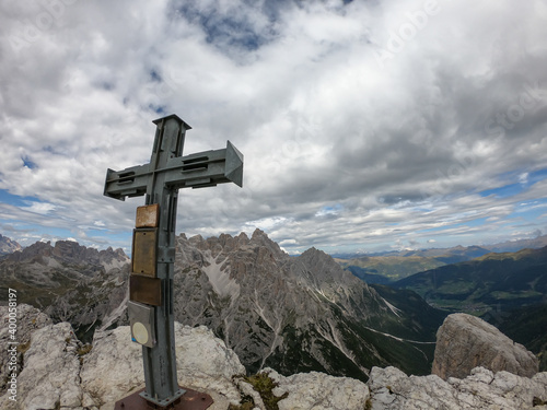 A wooden cross on high and desolated mountain peak in Italian Dolomites. In the back there are endless mountain chains. Raw and unspoiled landscape. Few clouds above the peaks. Sharp and steep slopes.