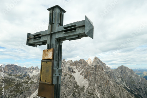 A wooden cross on high and desolated mountain peak in Italian Dolomites. In the back there are endless mountain chains. Raw and unspoiled landscape. Few clouds above the peaks. Sharp and steep slopes.