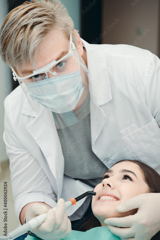 Blond man dentist in protective mask and white robe performs brushing of lady's teeth Blond man dentist in protective mask and white robe performs brushing of lady's teeth