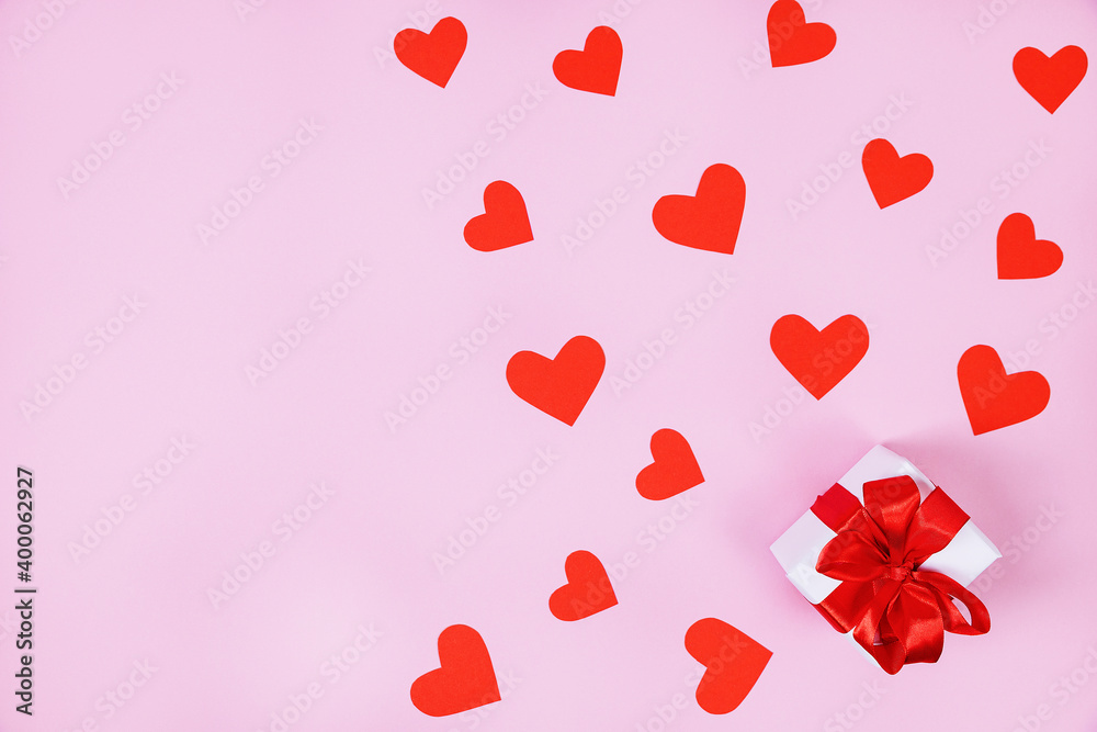Gift box with red ribbon, around lie red paper hearts. Pink background. The concept of Valentine's Day, birthday. Flat lay, top view, copy space.