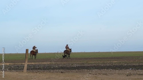 Two cowboys riding in the middle of the field, in slow motion, with their dogs by their side. photo