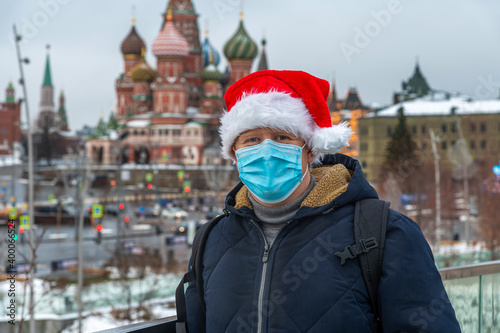 Portrait of russian man wearing blue protective face mask and red santa hat. Blurred Saint Basil's Cathedral and Moscow Kremlin in the background. Theme of new year in Russia during coronavirus.